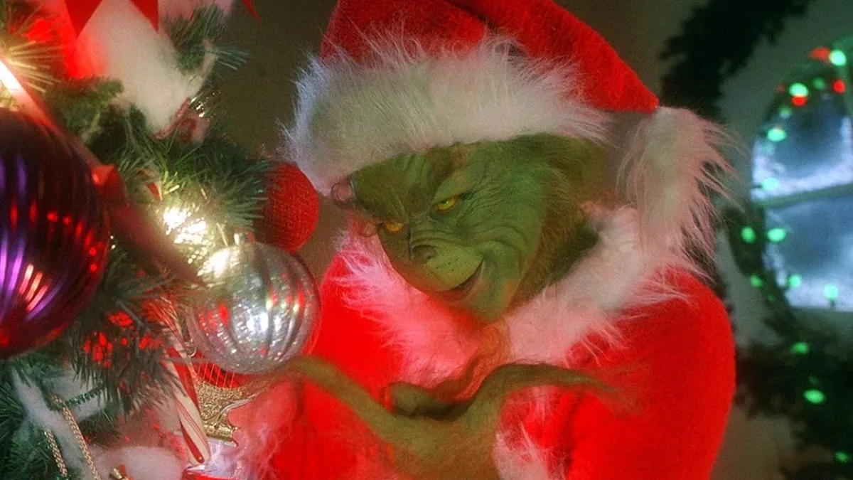 Jim Carrey as The Grinch in Santa hat, looking at ornament on tree
