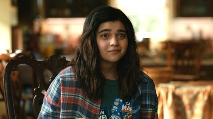 Image of Iman Vellani as Kamala Khan in 'The Marvels.' She is a brown Pakistani teenage girl with long, dark hair wearing a red and blue flannel shirt over a graphic tee depicting Avengers comics covers. She's sitting in a chair in her living room with a hopeful look on her face.