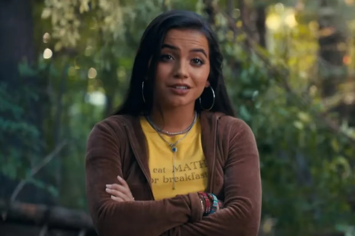 Screencap of Isabela Merced as Anya Corazon in the trailer for 'Madame Web.' She is a teenage Latina with long dark hair wearing silver hoop earrings and two silver necklaces over a yellow t-shirt that says "I eat MATH for breakfast" that she's wearing under a brown velvet hoodie. She's standing with her arms folded in a wooded area.