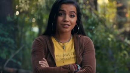 Screencap of Isabela Merced as Anya Corazon in the trailer for 'Madame Web.' She is a teenage Latina with long dark hair wearing silver hoop earrings and two silver necklaces over a yellow t-shirt that says 