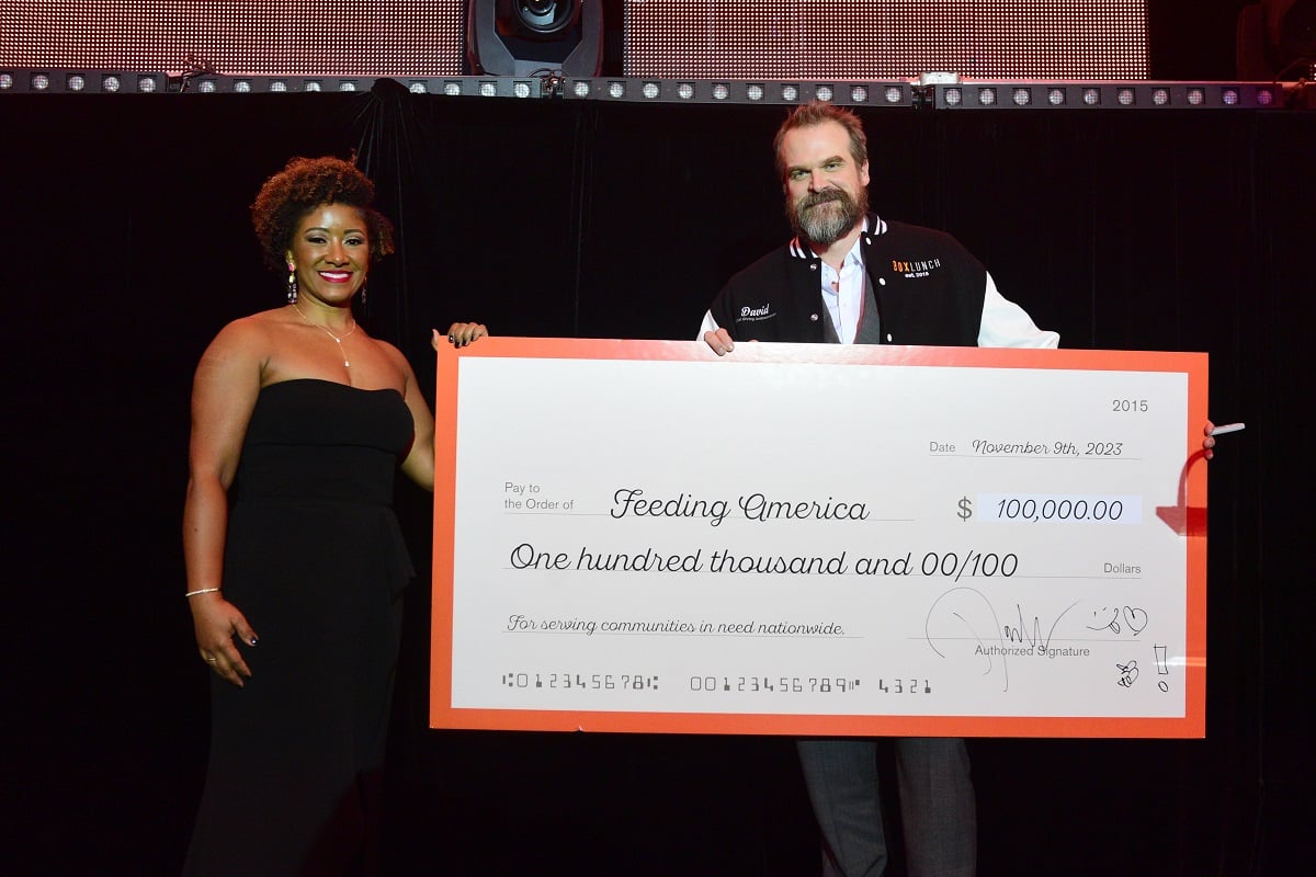 Image of Jennifer Polk and David Harbour standing together on stage holding a large fake $100,000 check from Harbour to Feeding America. Polk is a Black woman with short, curly dark hair wearing a strapless black jumpsuit, dangly earrings, and a thin diamond necklace. Harbour is a white man with short brown hair and a salt-and-pepper beard wearing a BoxLunch letterman jacket over a white button down and grey suit pants.