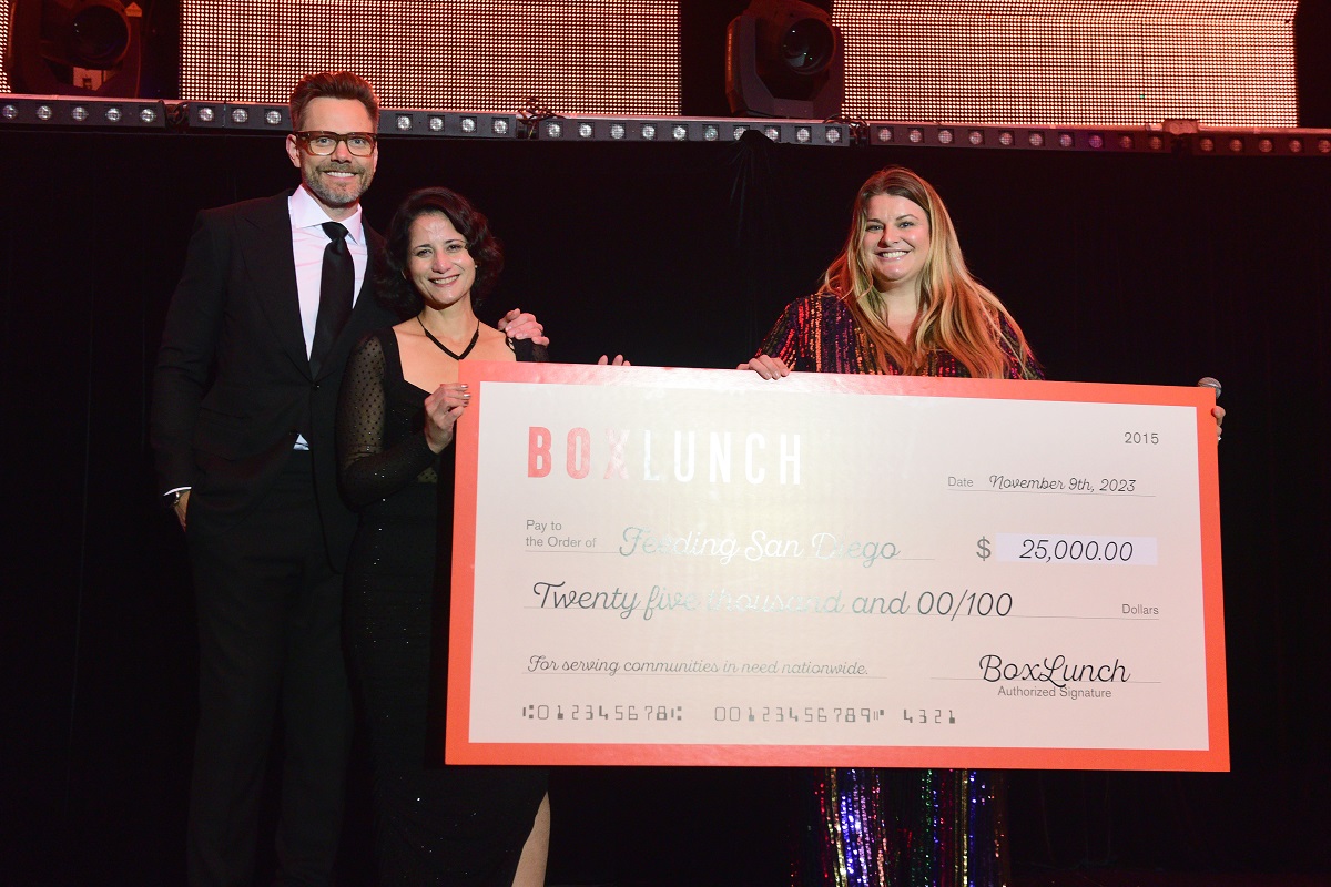 Image of Joel McHale, Erika Duran, and Ali Colbran standing together on stage holding a large fake $25,000 check from BoxLunch to Feeding San Diego. Joel McHale is a white man with a salt-and-pepper beard and glasses wearing a black suit and tie with a white shirt. Erika is a white woman with a chin-length bob wearing a black dress. Ali is a fat white woman with long blonde hair wearing a glittery, rainbow jumpsuit. 