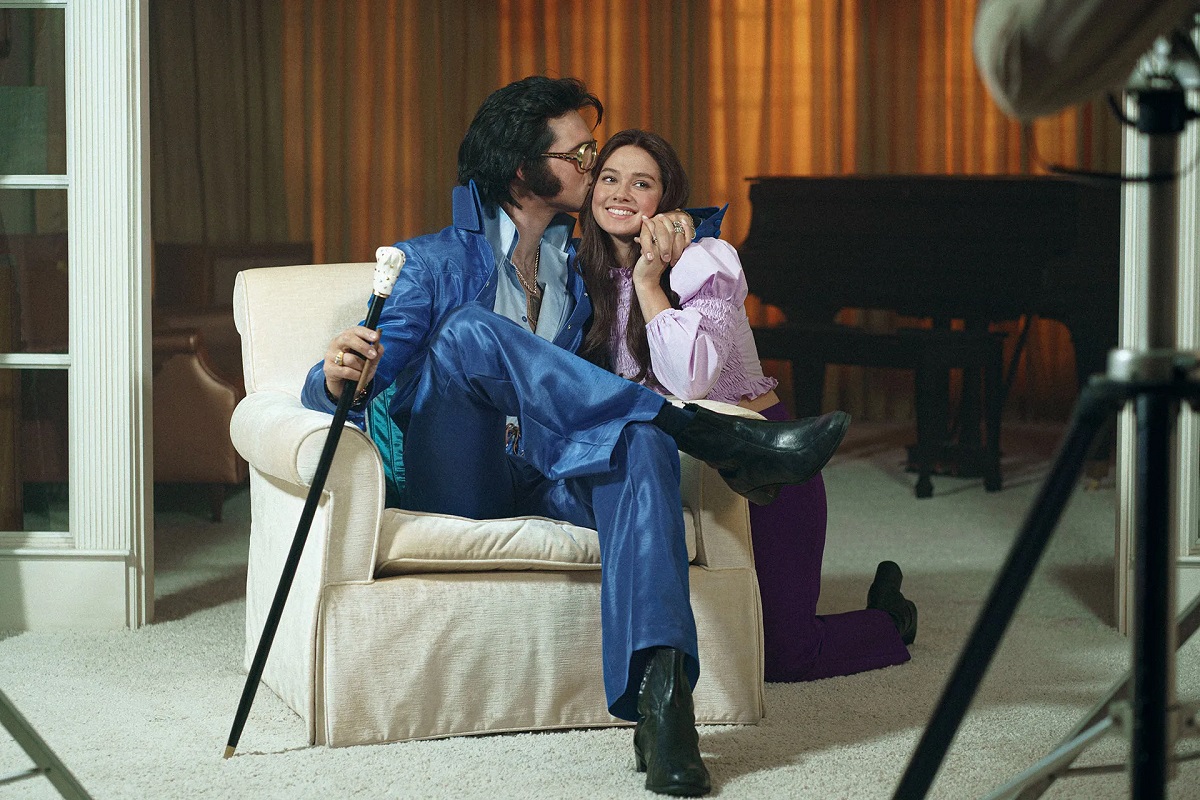 Image of Jacob Elordi as Elvis Presley and Cailee Spaeny as Priscilla Beaulieu in the film 'Priscilla.' Elvis is a white man with shaggy black hair and mutton chop sideburns wearing large sunglasses, a blue suede jumpsuit over a lighter blue buttondown shirt with the first few buttons opened, and silver dogtags around his neck. He's seated in a white armchair holding a walking stick in one hand with his other arm around Priscilla, who's kneeling beside the chair. He's kissing the side of her face as she looks forward, smiling. Priscilla is a white young woman with long dark hair wearing a pink blouse with puffy long sleeves and black pants. 