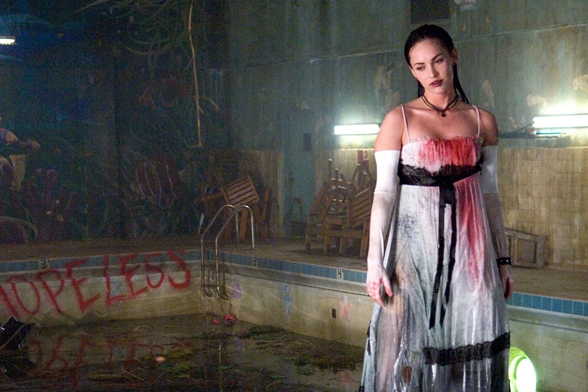 Image of Megan Fox as Jennifer in a scene from 'Jennifer's Body.' She is a white teen girl with long black hair hovering over a dilapidated swimming pool that's filled with algae and has the word "Hopeless" written in red spray paint on one end behind her. She's all wet and wearing a black and white prom dress that's covered in blood.   