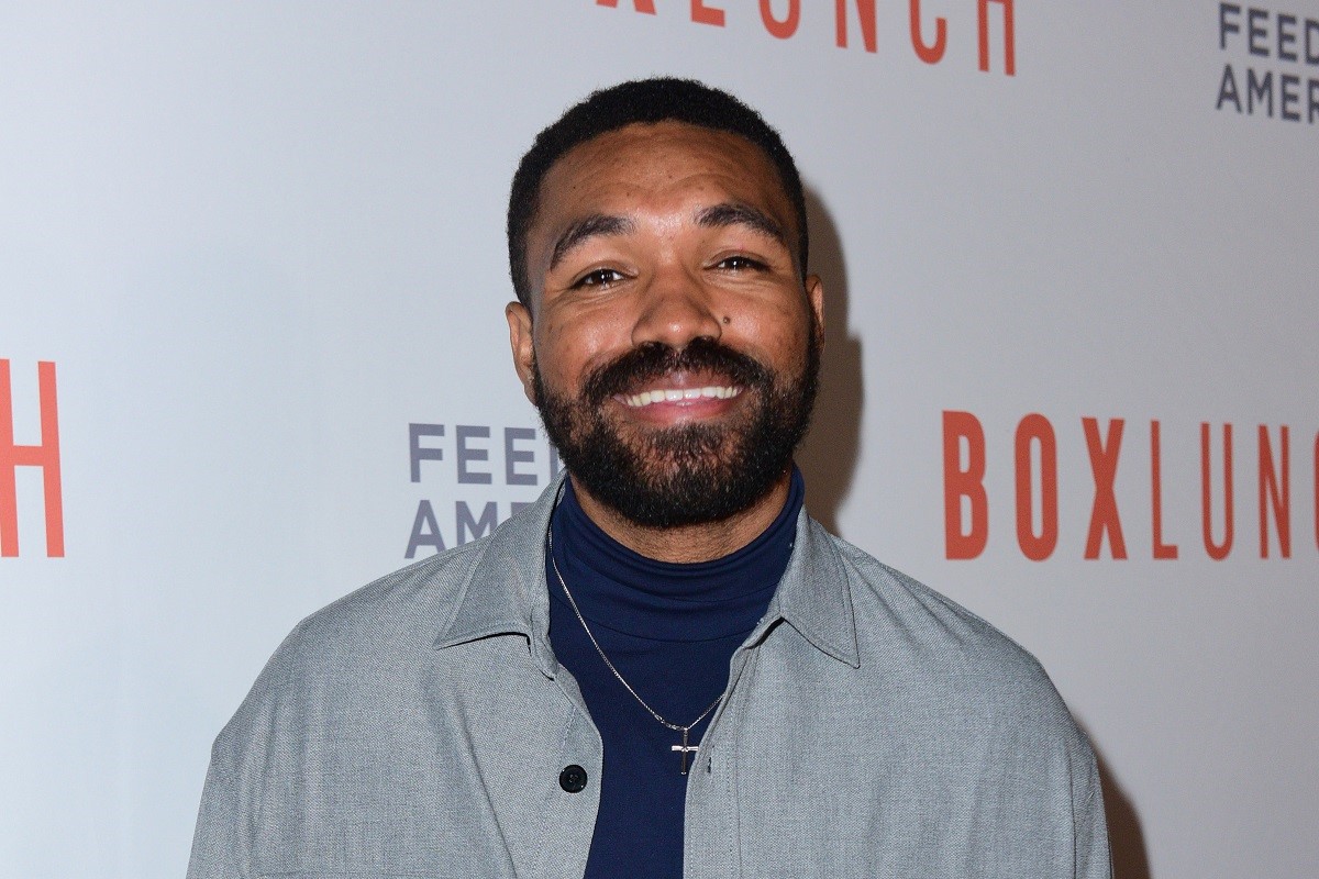Image of Jordan Howlett on the red carpet at the BoxLunch Holiday Gala 2023. He is a Black man with short black hair and a black beard. He's wearing a grey jacket over a navy blue turtleneck with a silver cross pendant around his neck. He's smiling at the camera and standing in front of a step and repeat that has the BoxLunch and Feeding America logos on it.