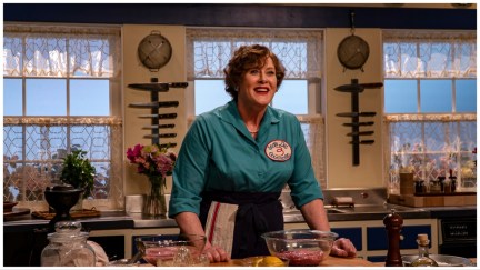 Julia Child (Sarah Lancashire) wears a blue shirt and skirt and stands in her kitchen hosting her cooking show.
