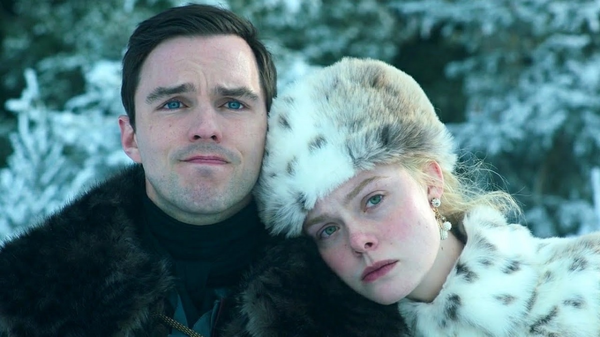 Image of Nicholas Hoult as Peter and Elle Fanning as Catherine in a scene from Hulu's 'The Great.' Peter is a white man with short, dark hair wearing a black fur coat. Catherine is a white woman with blonde hair wearing a brown and white fur hat and a matching fur coat. They are sitting outside in the snow in front of trees, and Catherine is resting her head on Peter's shoulder as they both look out into the distance. 