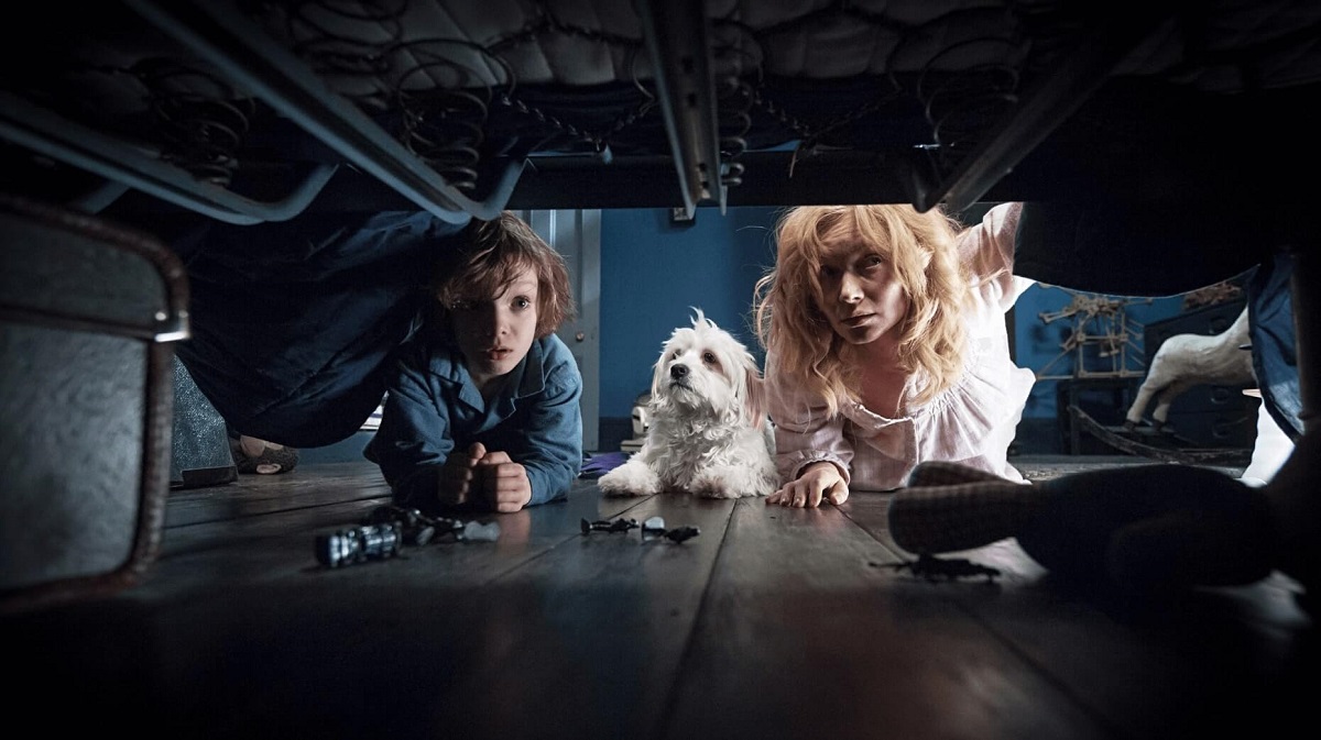 Image of Noah Wiseman (Samuel) and Essie Davis (Amelia) in a scene from 'The Babadook.' They are both on the floor looking under a bed with their white dog between them. Samuel is a white young boy with shaggy brown hair wearing blue pajamas. Amelia is a white blonde woman wearing a white long-sleeved nightgown.