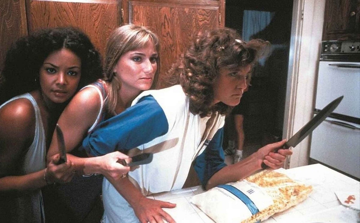 Andree Honore (Jackie), Debra De Liso (Kim) and Michele Michaels (Trish) in a scene from 'The Slumber Party Massacre.' Jackie is a Black teen girl with long, natural hair wearing a white sleeveless nightgown. Kim is a blonde teen girl in a tank top. Trish is a white teen girl with curly brown hair wearing a jersey with blue sleeves. They are standing huddled together in a kitchen all holding large knives.