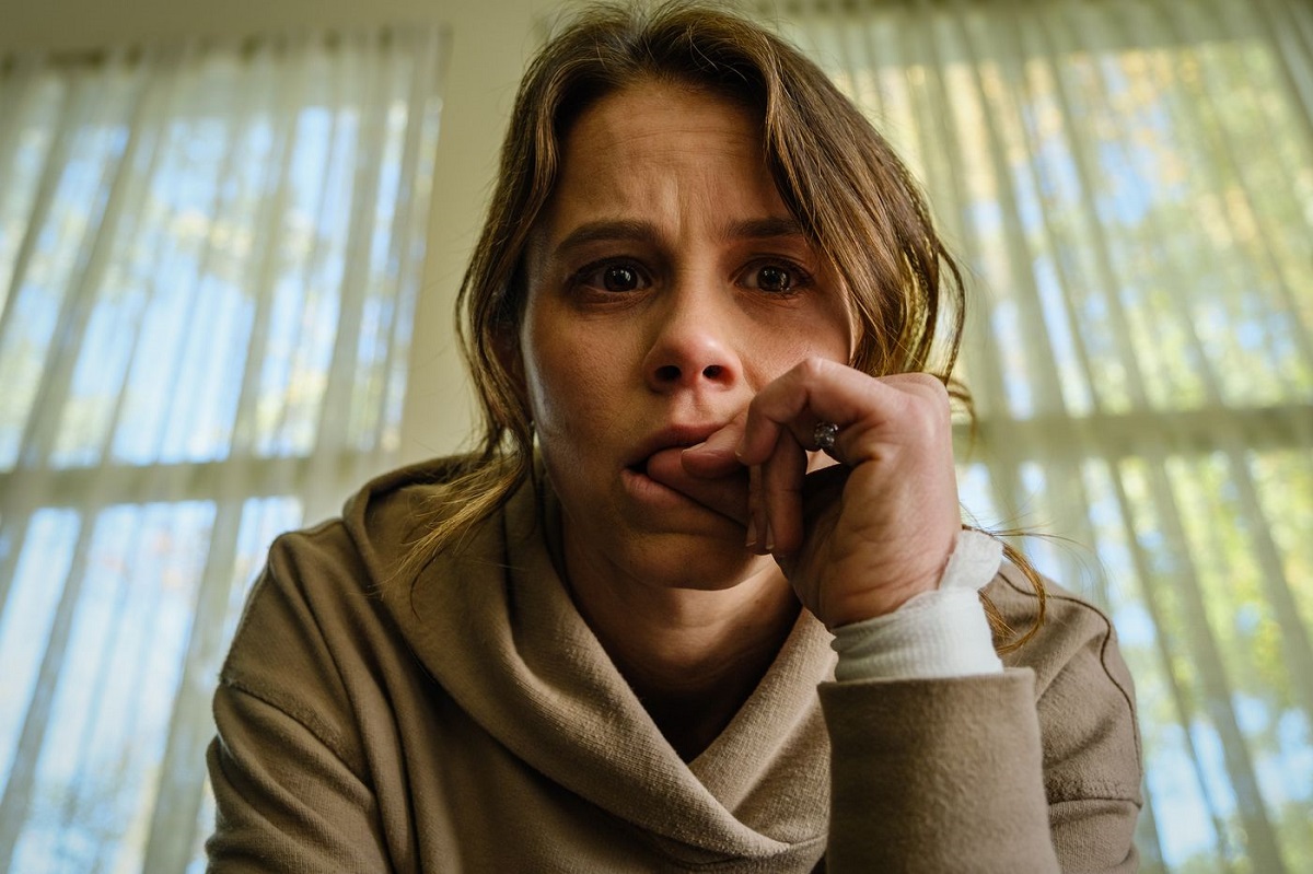 Sosie Bacon (Rose) in a scene from 'Smile.' She is a white woman with her brown hair in a ponytail with tendrils hanging in her face. She's wearing a beige hooded sweatshirt and seated on a couch in front of a window biting her thumbnail and looking nervous.