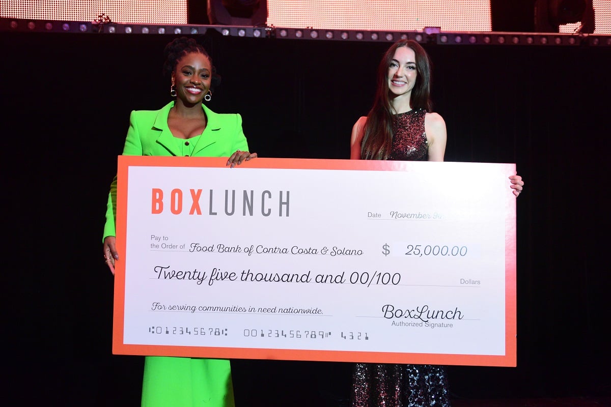 Image of Teyonah Parris and Cassidie Bates of Food Bank of Contra Costa and Solano standing together on stage holding a large fake $25,000 check from BoxLunch to Food Bank of Contra Costa and Solano. Teyonah is a Black woman with her black hair in tight braids wearing a bright green suit. Cassidie is a white woman with long brown hair wearing a sparkly black and red sleeveless dress. 