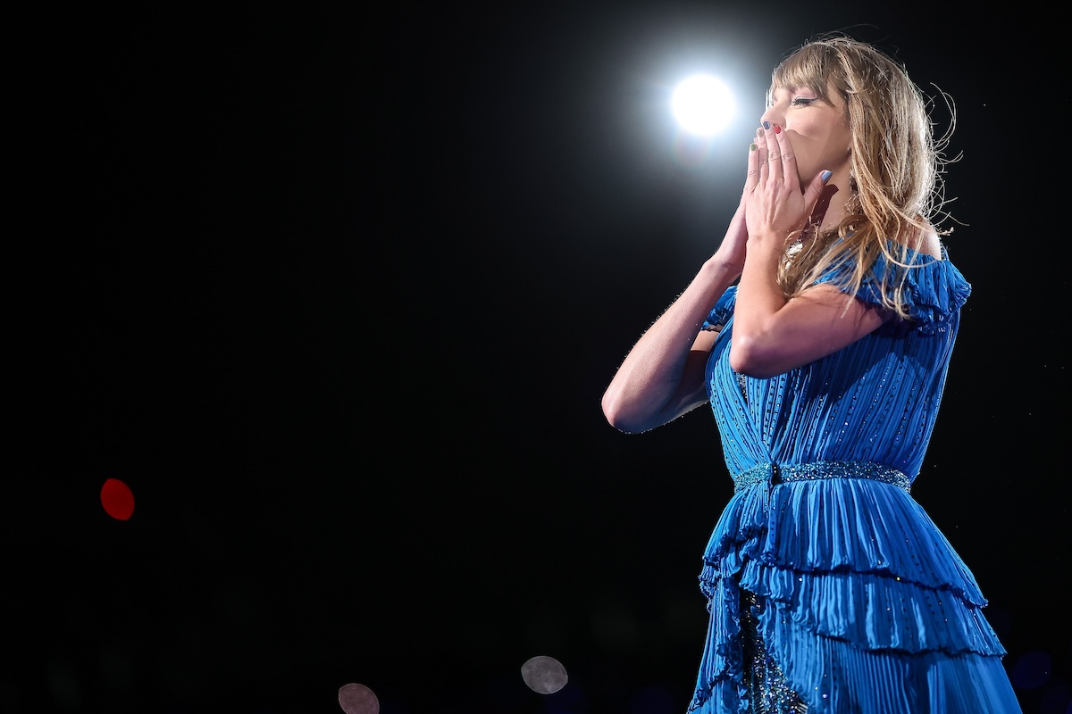 Taylor Swift in a blue dress on stage performing, holding her hands to her mouth to blow a kiss to the audience.