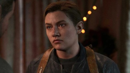 Image of Abby Anderson in a cutscene from the video game 'The Last of Us, Part 2.' Abby is a white teenage girl with brown hair in a ponytail wearing a black jacket over a black t-shirt with a brown backpack on her back. She is looking at someone with a serious expression.