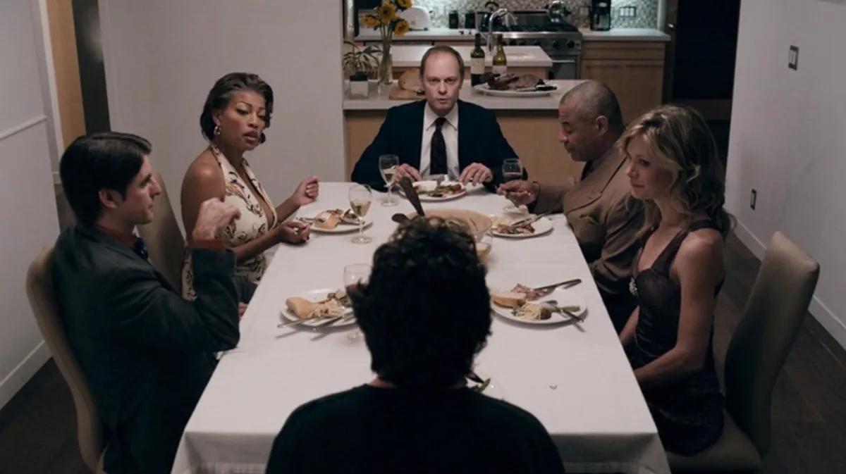 a group of people sit around a dinner table looking at a mystery guest who has his back to the camera. 'The Perfect Host.'