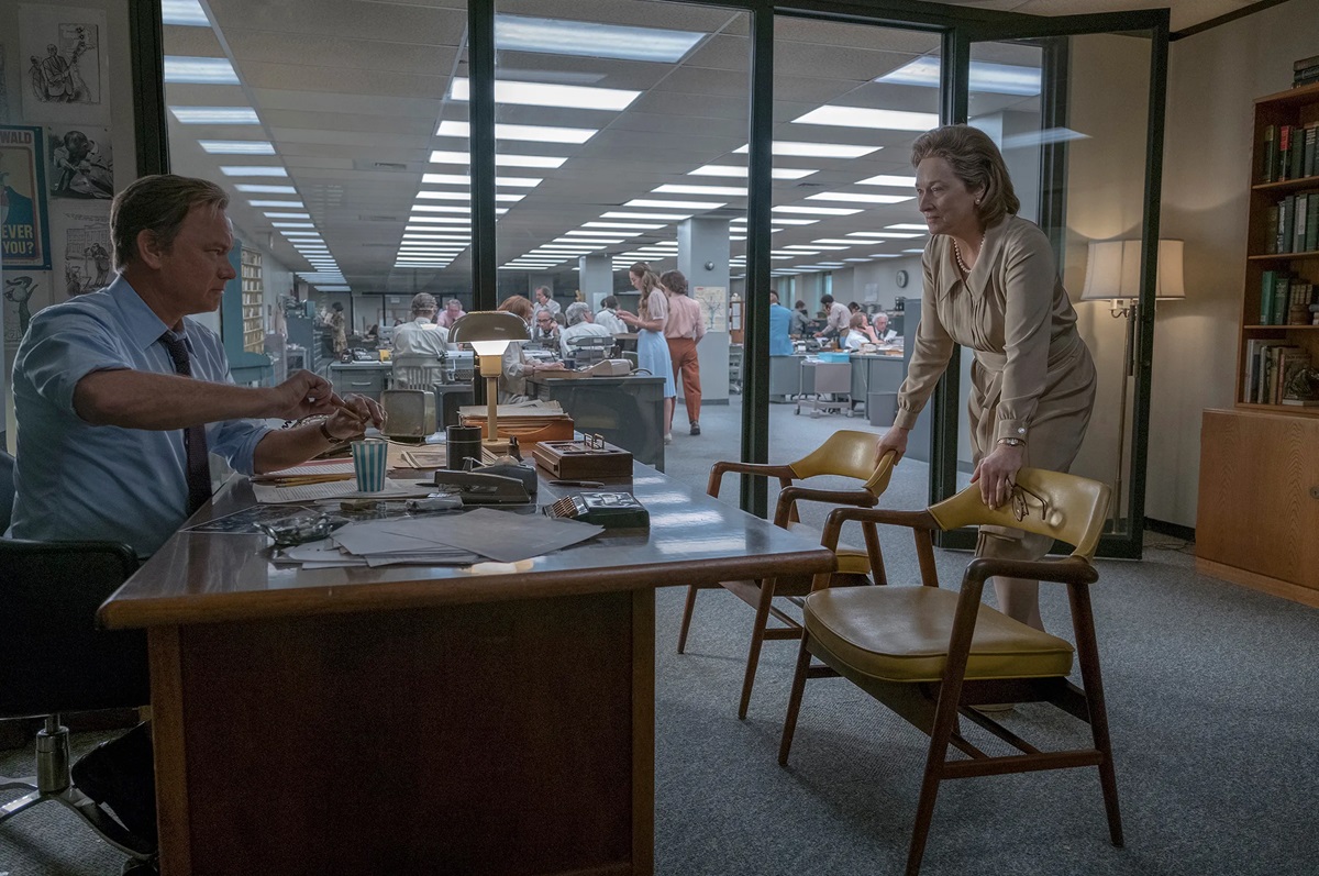 man sits behind desk while woman stands with hands on a chair, looking at him