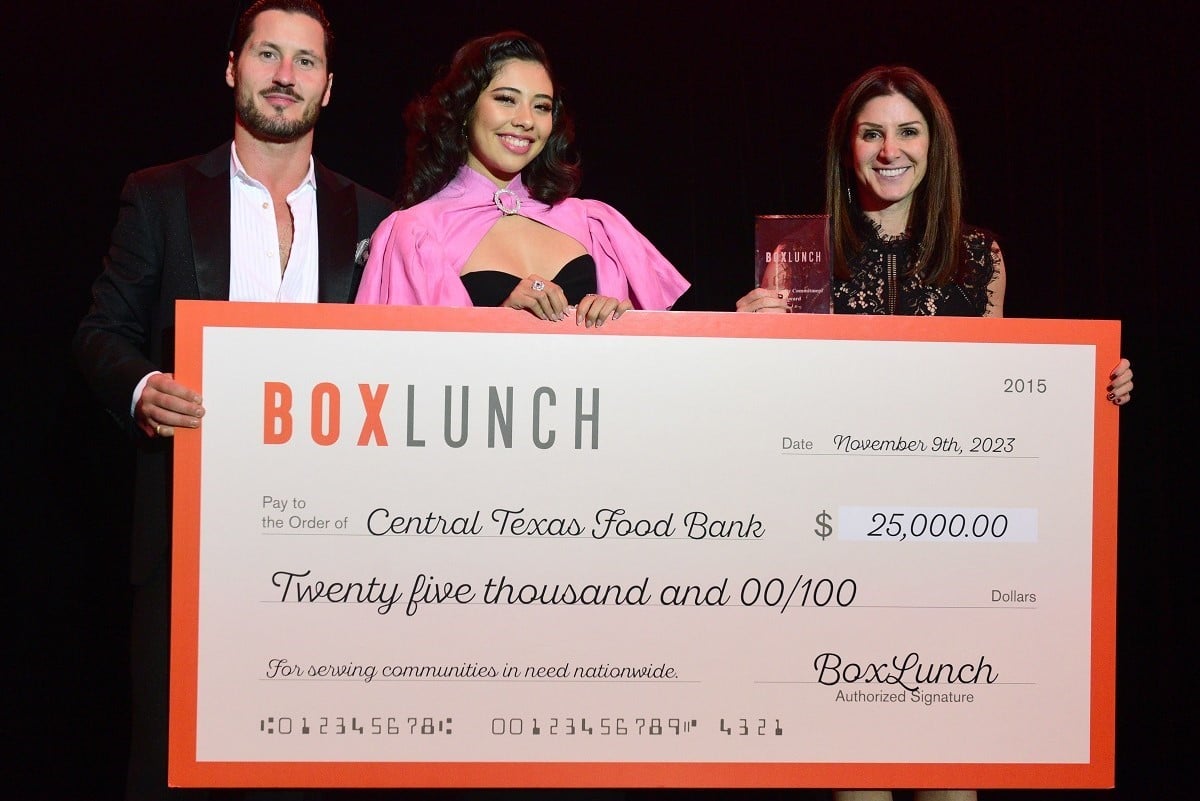 Image of Val Chmerkovskiy, Xochitl Gomez, and Sari Vatske standing together on stage holding a large fake $25,000 check from BoxLunch to Central Texas Food Bank. Val is a white man with short dark hair and a thin dark beard wearing a black suit and a white buttondown with the first three buttons undone. Xochitl is a Latina teenage girl wearing a black dress with a hot pink shawl with short sleeves and a round diamond clasp at the neck. Sari is a white woman with shoulder-length brown hair wearing a black dress and holding an award in her hand. 