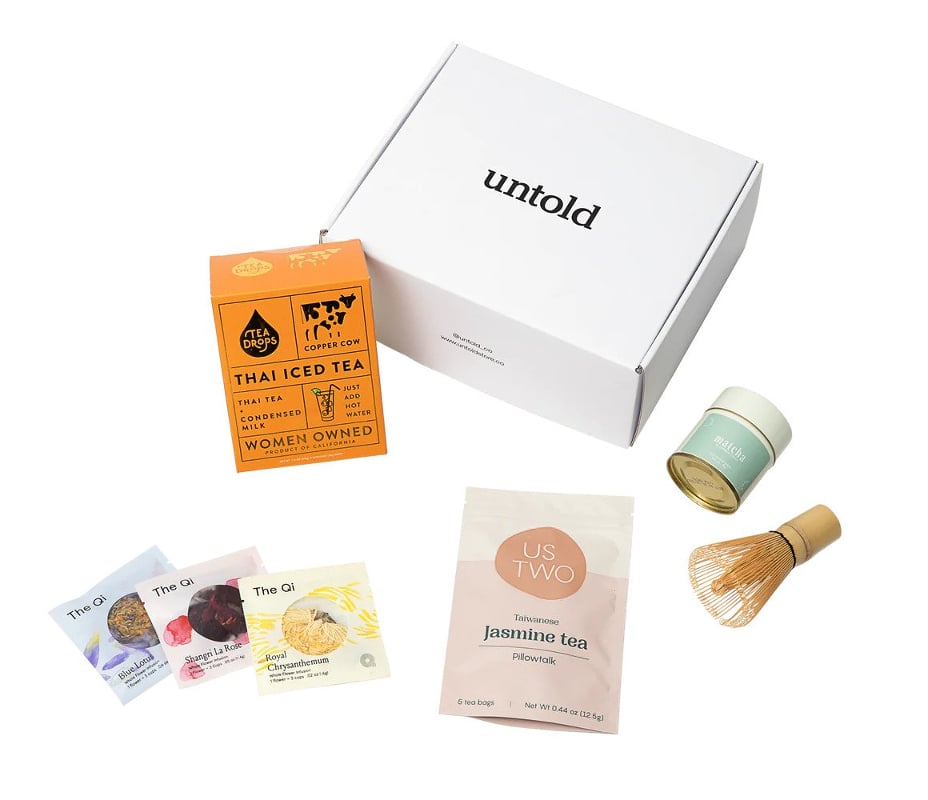 A white cardboard box with a selection of different packages of tea and a matcha whisk in front of it.
