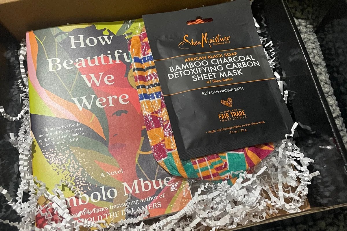 A box with white paper packaging strips; inside it is a copy of "How Beautiful We Were" by Imbolo Mbue and a black packet with the label Bamboo Charcoal Facemask on it.