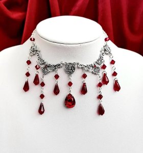 A silver necklace made of swirls and roses with red crystal beads and drops coming down from it.