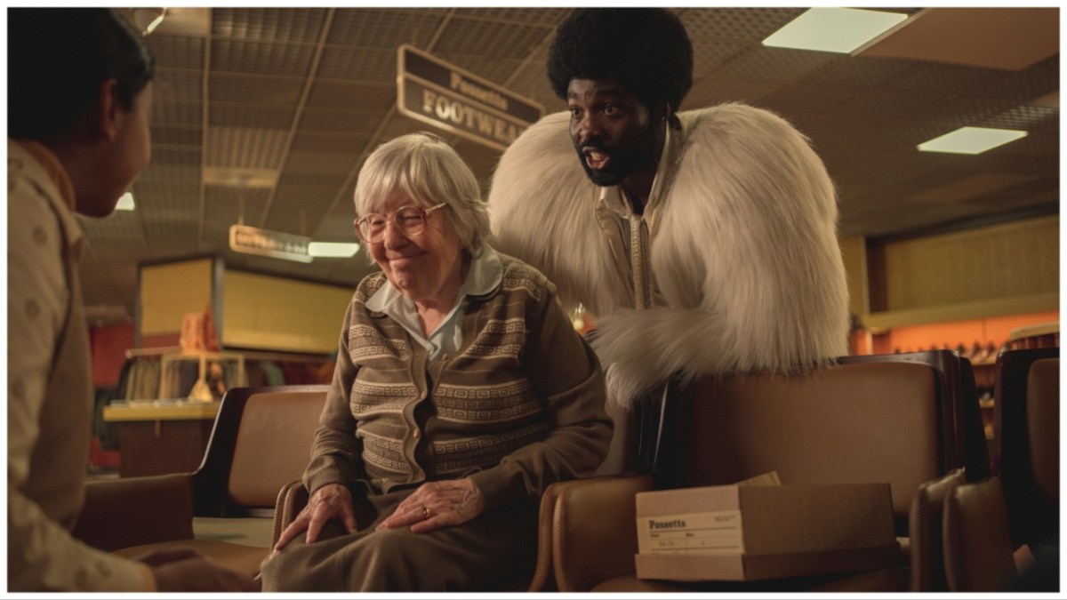 A Black man in a white fluffy coat stands behind an old woman trying on shoes.
