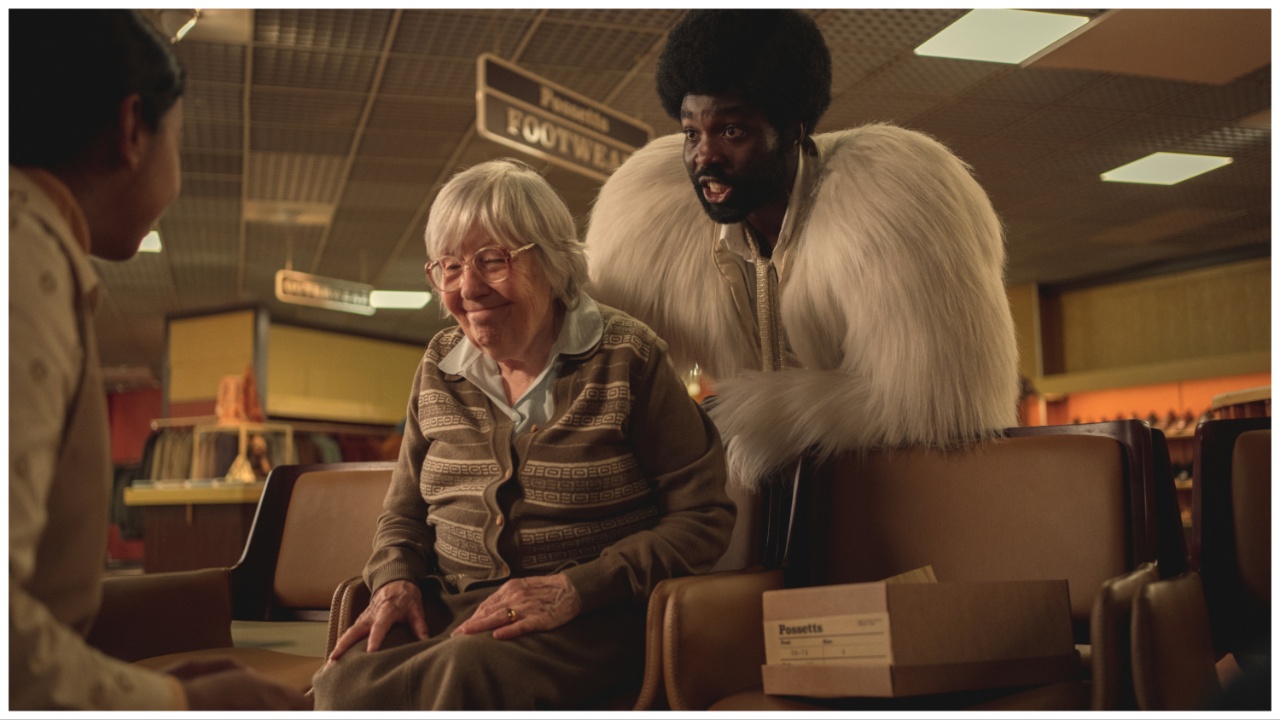 A Black man in a white fluffy coat stands behind an old woman trying on shoes.