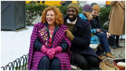 Flora the Genie (Melissa McCarthy) wears a purple coat and sits on a bench next to Bernard (Paapa Essiedu) who is wearing a winter hat and black jacket.