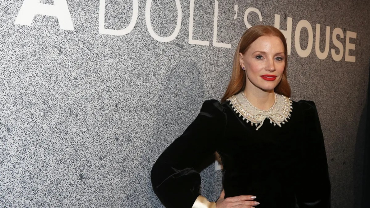 Jessica Chastain, in a black dress with a white collar, stands in front of a sign for 'A Doll's House'. 
