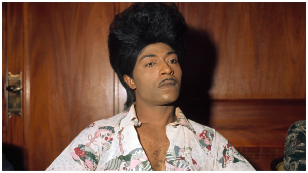 Little Richard, a Black man with a bouffant hairdo and a patterned shirt against a wooden wall.