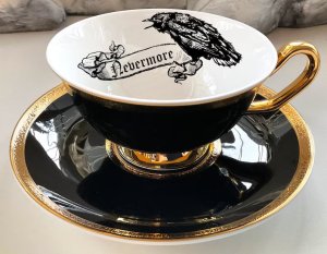 A black tea cup and saucer with gold handle and rims, and a line drawing of a raven and the word nevermore printed on the inside.