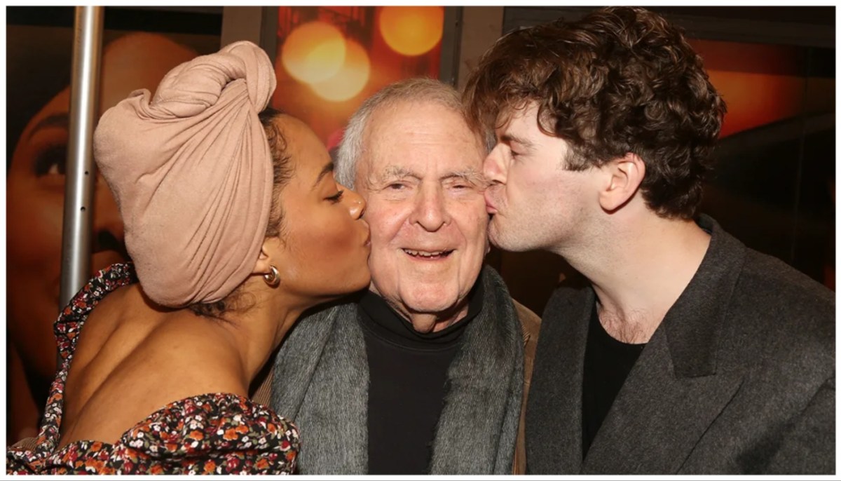 A woman in a head wrap and a brunette man stand on either side of an older man, each kissing him on the cheek. 