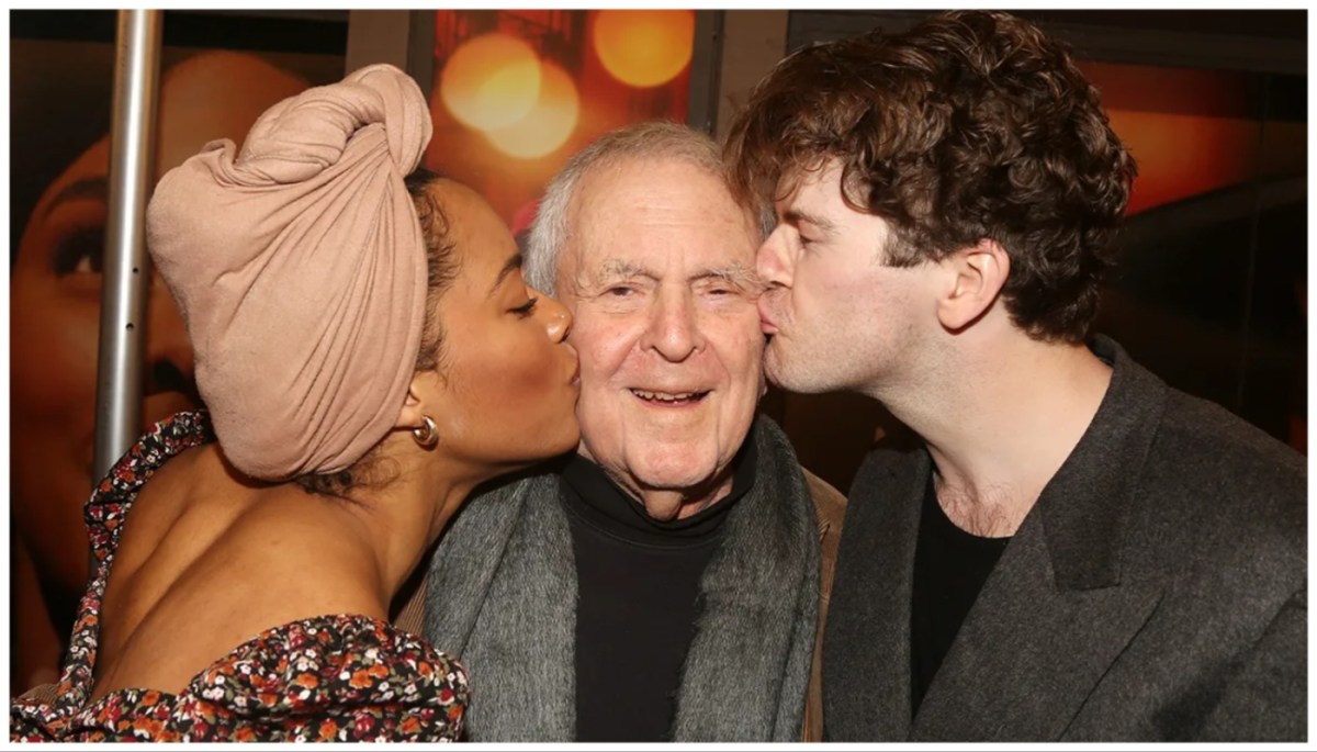 A woman in a head wrap and a brunette man stand on either side of an older man, each kissing him on the cheek. 