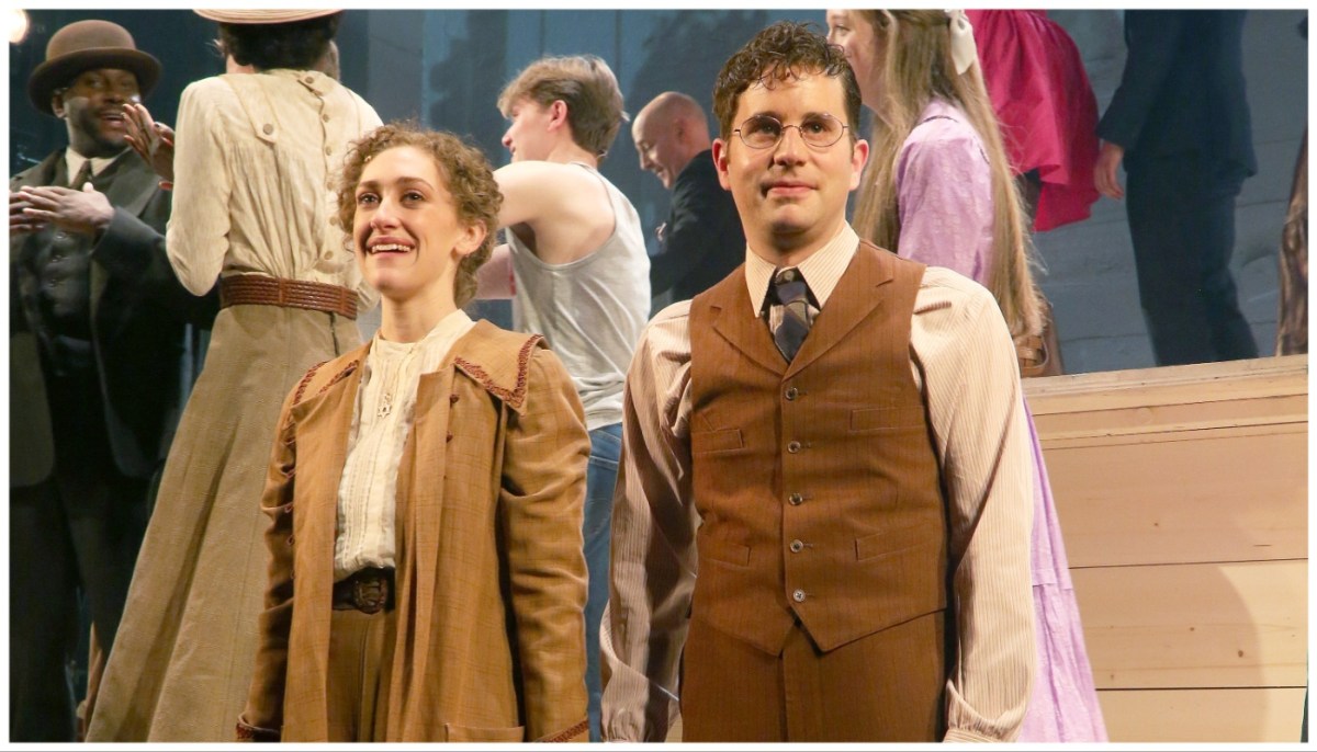 A woman  and a man stand side by ide holding hands as people walk behind them. They are dressed in clothing from the 1910s.