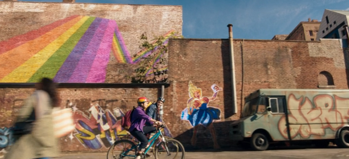Kamala and Bruno ride their bikes past a brick building with Captain Marvel in a princess gown painted on it.