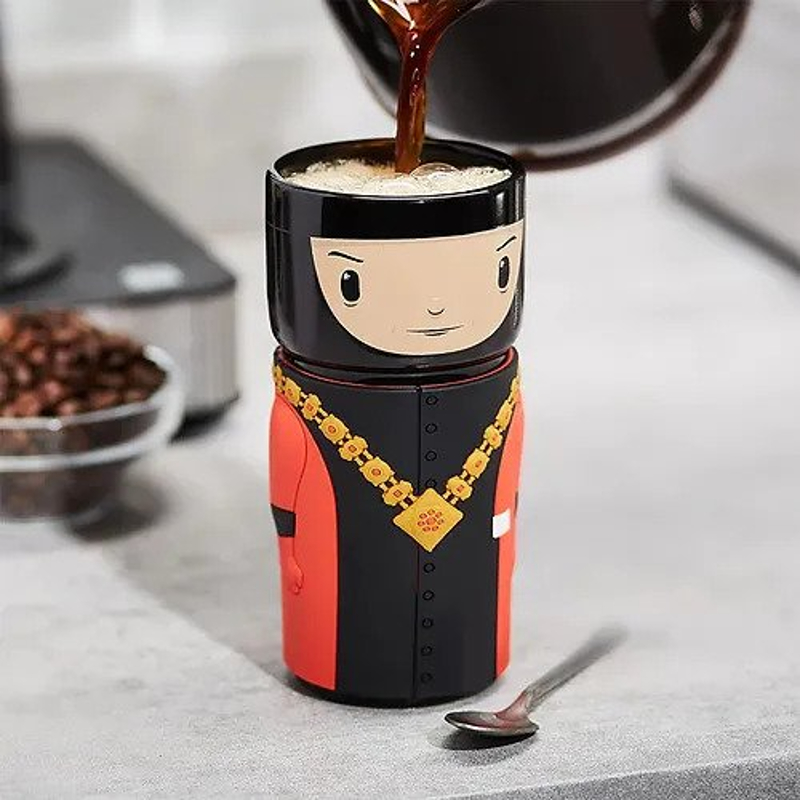 A cylindrical cup decorated to look like Q in a cartoonish fashion. He's wearing his red and black robes and gold chain and is having coffee poured into him.