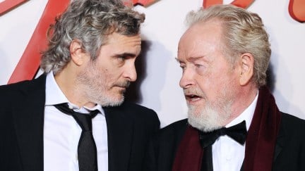 Joaquin Phoenix and Ridley Scott on the Napoleon red carpet, leaning into each other and talking close.