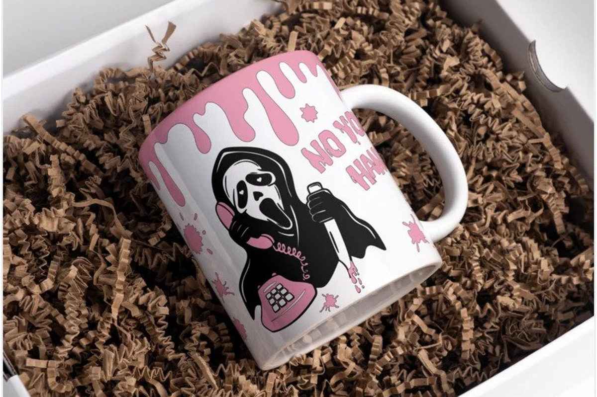 A white mug with pink blood spatter and the ghost face killer in black and white on it. The test "no you hang up" is partially visible and the mug is in a box full of brown shredded paper.