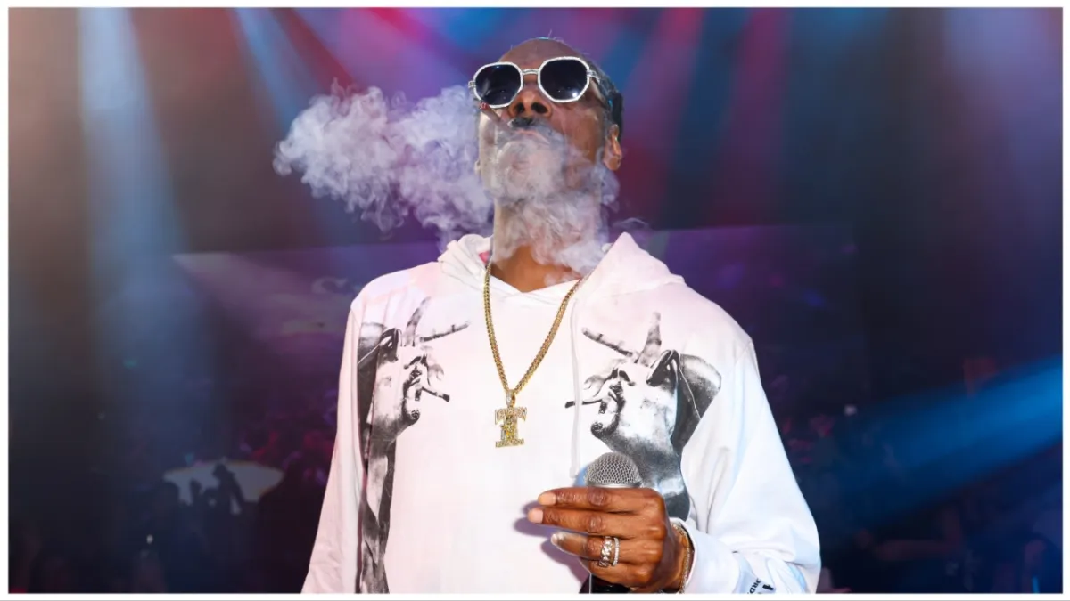 Snoop Dogg stands onstage with a joint in one hand and a microphone in the other in an all-white track suit.