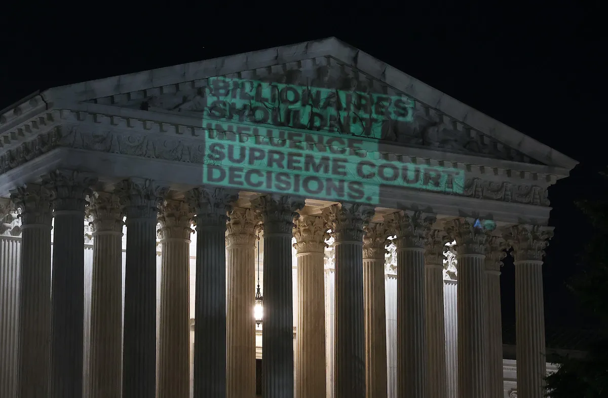 A graphic reading "billionaires shouldn't influence supreme court decisions" is projected onto the supreme court building at night.