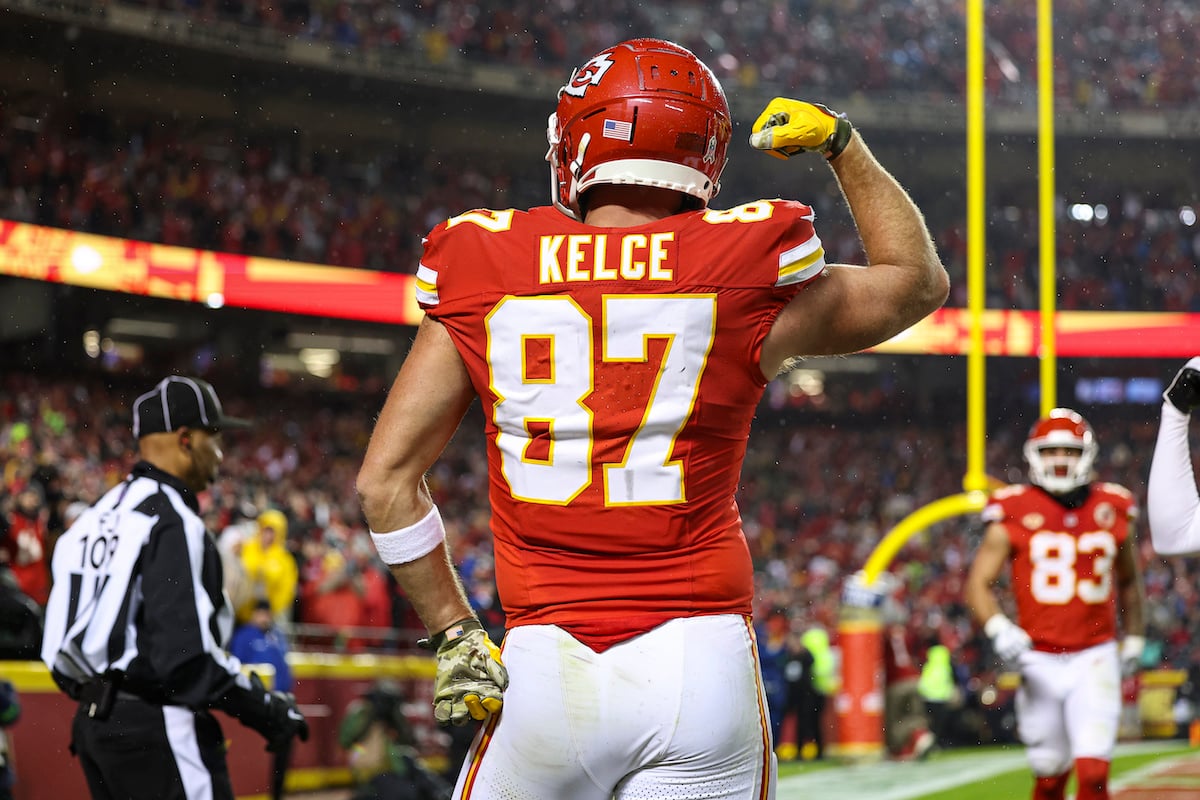 Travis Kelce, seen from behind on the field during a football game, posing with one hand on his hip and the other arm flexing his bicep.