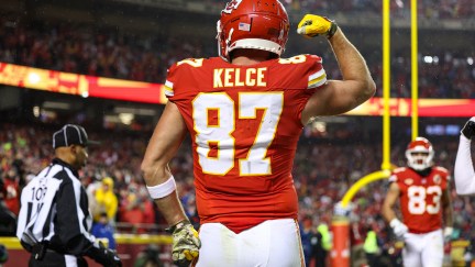 Travis Kelce, seen from behind on the field during a football game, posing with one hand on his hip and the other arm flexing his bicep.