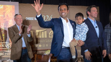 Vivek Ramaswamy holds his son and waves to attendees at an event. Ron DeSantis stands next to him doing his robotic human-like smile.