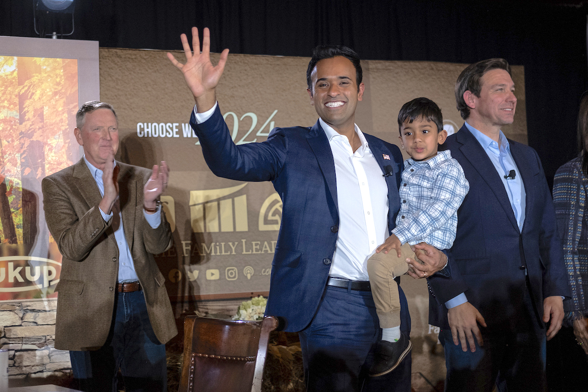 Vivek Ramaswamy holds his son and waves to attendees at an event. Ron DeSantis stands next to him doing his robotic human-like smile.