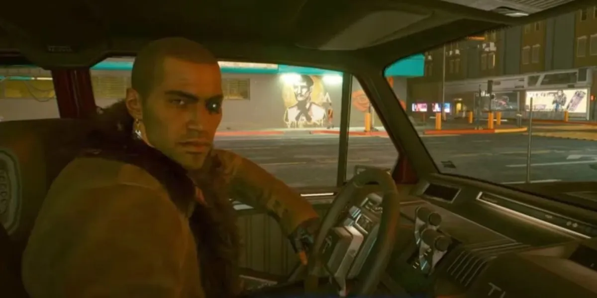 A man driving a car looks over at the camera in the passenger seat in "Cyberpunk 2077"