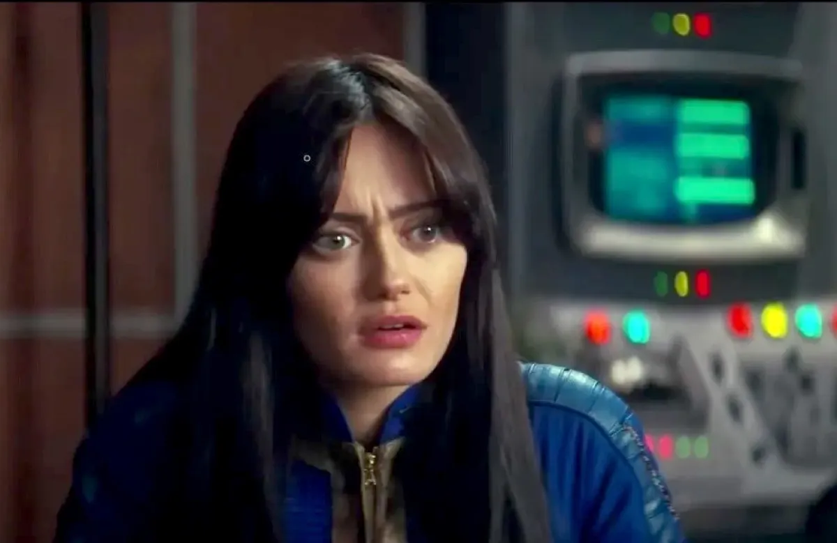 Ella Purnell as Lucy in a scene from Amazon's 'Fallout' series. She is a young white woman with long dark hair and long bangs parted down the middle. She's wearing a blue 'Fallout' vault suit and sitting in a room with computer consoles behind her. She has a confused expression on her face.