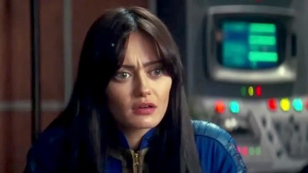 Ella Purnell as Lucy in a scene from Amazon's 'Fallout' series. She is a young white woman with long dark hair and long bangs parted down the middle. She's wearing a blue 'Fallout' vault suit and sitting in a room with computer consoles behind her. She has a confused expression on her face.