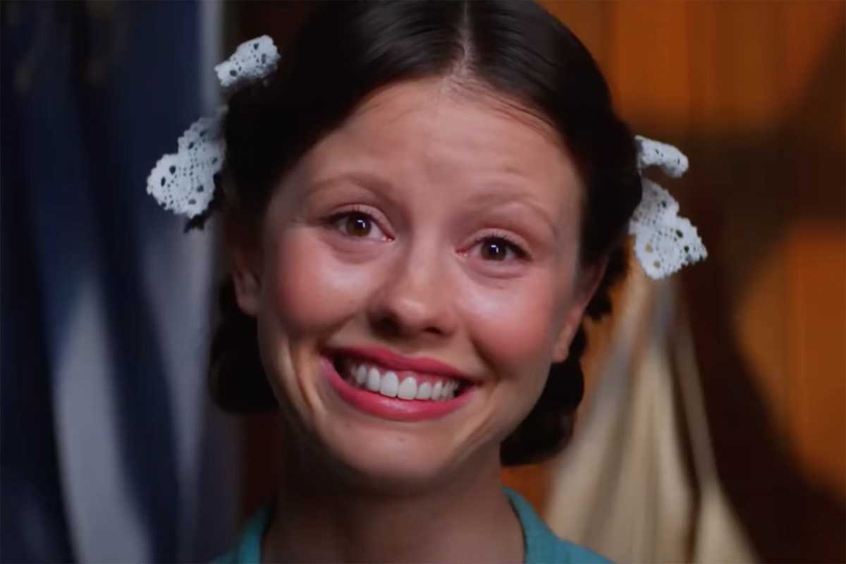 Mia Goth in a scene from the film 'Pearl.' She is a young white woman with a big smile and a blank stare staring into the camera. Her long dark hair is up in two braids looped up on either side of her head with white bows tying them up. 