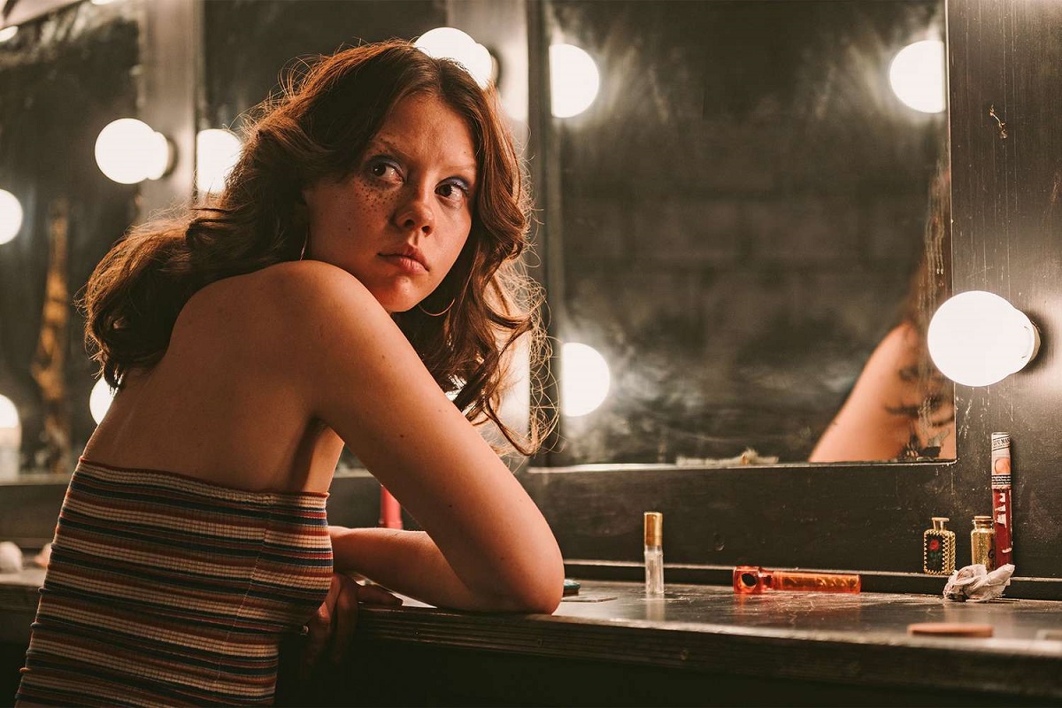 Mia Goth in a scene from the film 'X.' She is a young white woman seated at a make-up table with large bulbs around the mirror. Her arms are folded on the table as she looks over her shoulder. She has long, brown hair and is wearing a red, black, and white striped tube top. It's a 1970s period look. 