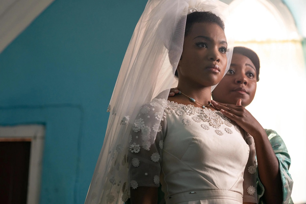 Image of Mia Isaac as young Eleanor in 'Black Cake.' She is a young Black woman in a wedding dress. Another young Black woman stands beside her fixing her necklace. They're standing in a room with light blue walls. 