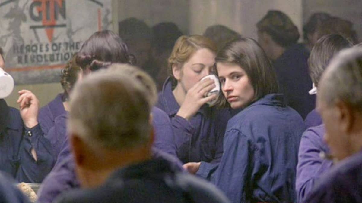 Susanna Hamilton as Julia in a scene from the film adaptation of '1984.' She is a young white woman with dark hair in a bob that's parted on the side and wearing dark blue coveralls seated at a cafeteria table surrounded by other white men and women in similar coveralls. While everyone else is eating and looking down, she's looking over her shoulder at the camera. 