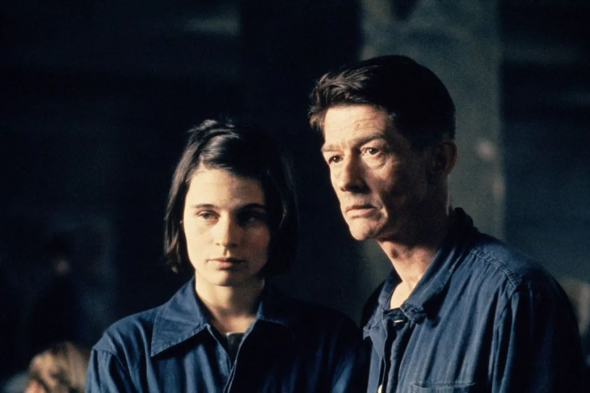 Susanna Hamilton as Julia and John Hurt as Winston in the film adaptation of '1984.' Julia is a young, white woman with dark hair in a bob parted on the side and wearing dark blue coveralls. Winston is a middle-aged white man with short, dark hair also wearing dark blue coveralls. They are standing next to each other and looking at something off camera. 
